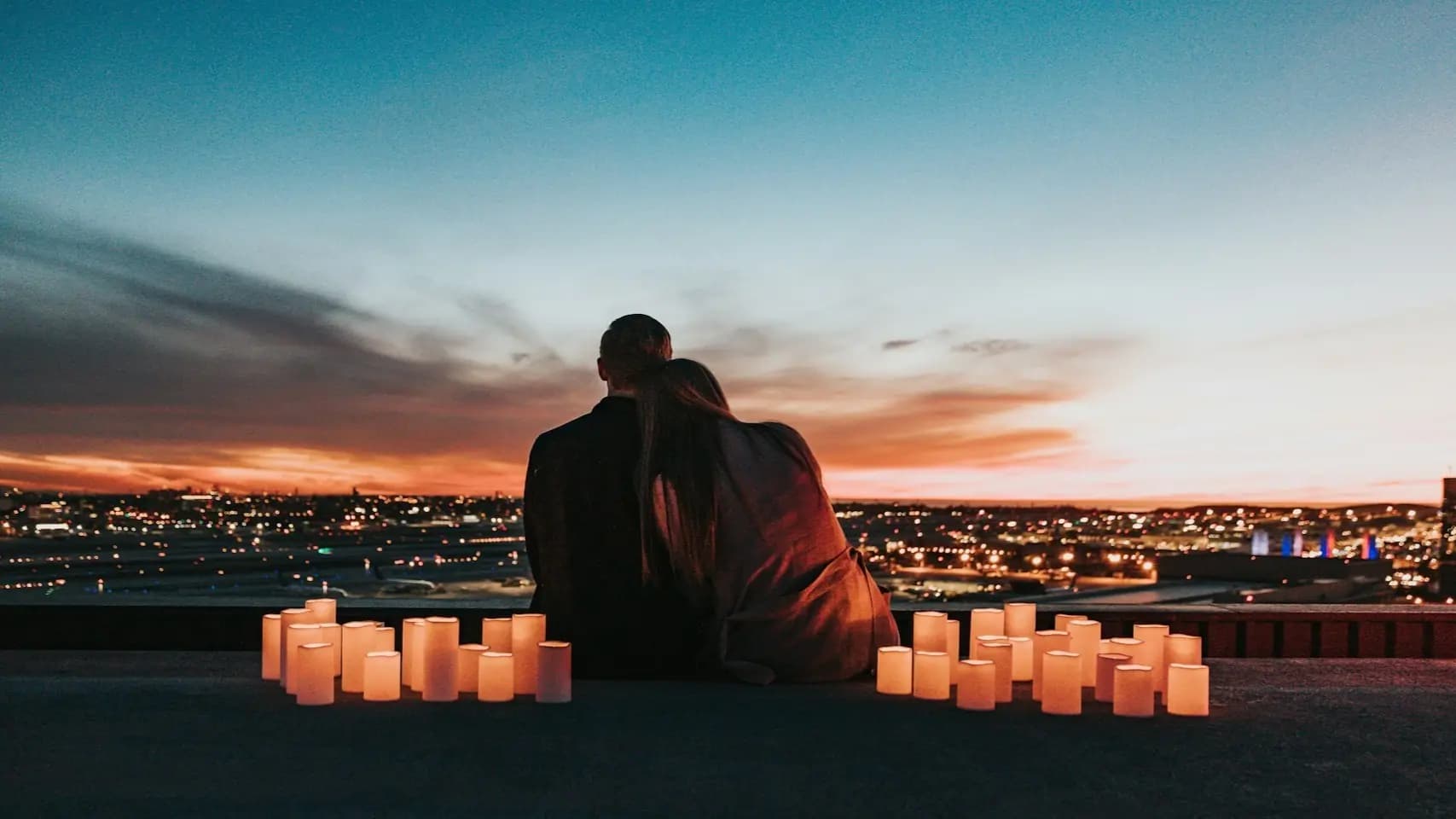 A couple sits close together on a rooftop at sunset, surrounded by glowing candles and overlooking city lights.